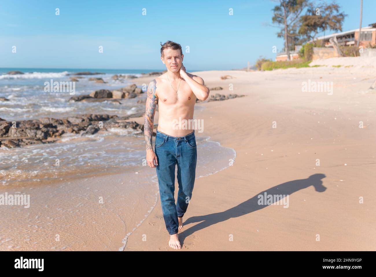 Attraente giovane caucasico nudo-chested uomo con braccio pieno tatuaggio che cammina sulla spiaggia a piedi nudi. Vista panoramica sull'oceano, foto mattutina, jeans. Foto Stock