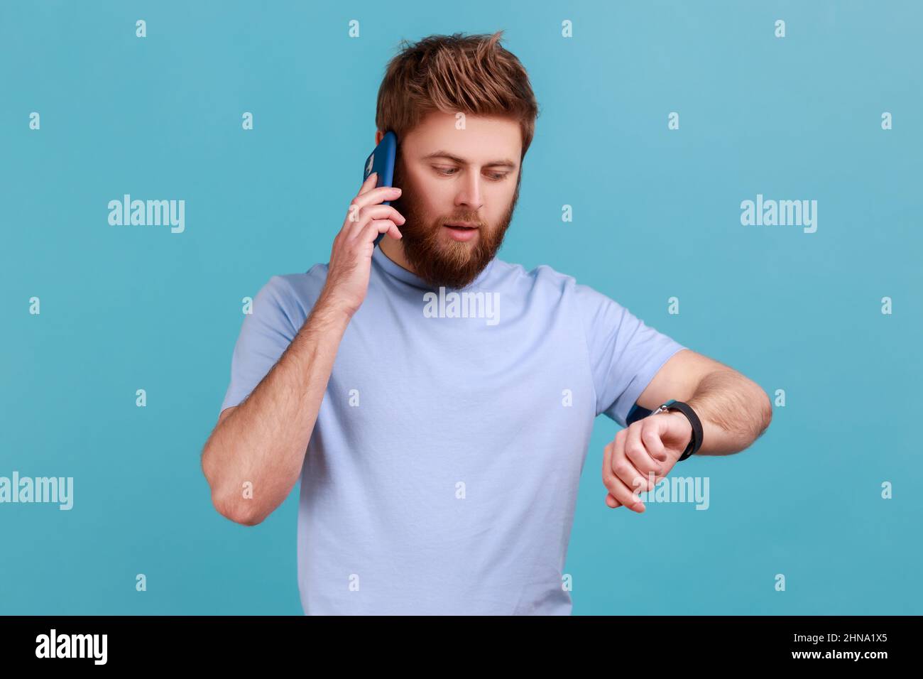 Ritratto di serio concentrato barbuto uomo parlare al telefono e guardare il tempo sui suoi orologi da polso, dicendo di essere in ritardo per l'incontro. Studio interno girato isolato su sfondo blu. Foto Stock