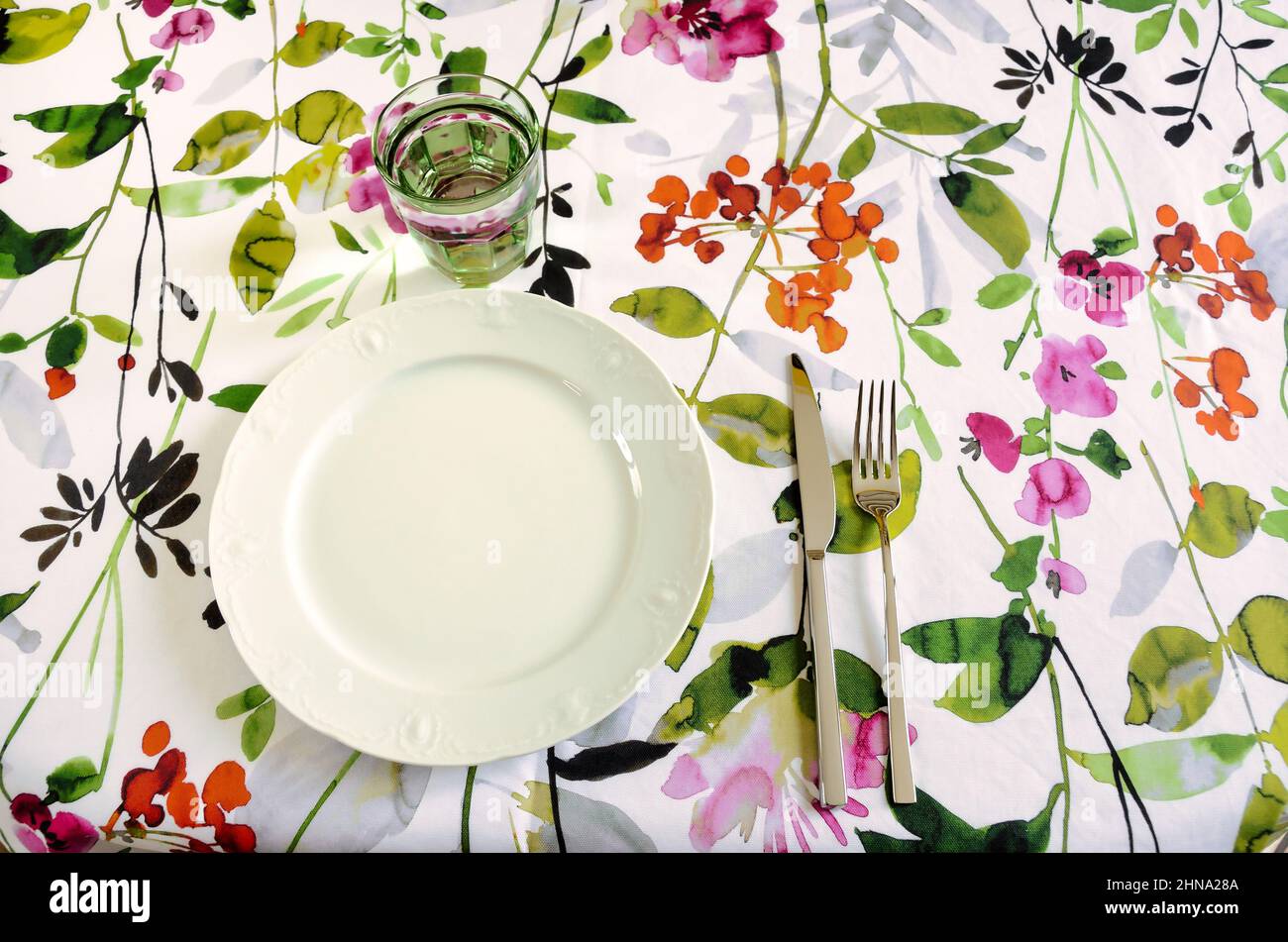 set da tavolo con tovaglia floreale, piatto bianco, vetro verde e posate in acciaio inox Foto Stock