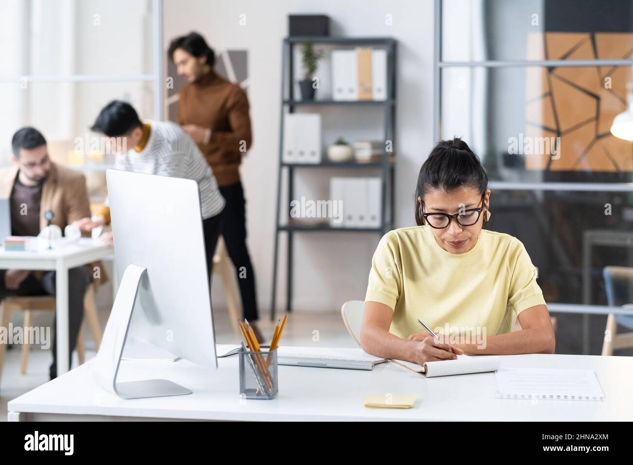 Giovane donna d'affari in occhiali seduti sul posto di lavoro con il computer e prendere appunti in notebook durante il suo lavoro in ufficio Foto Stock