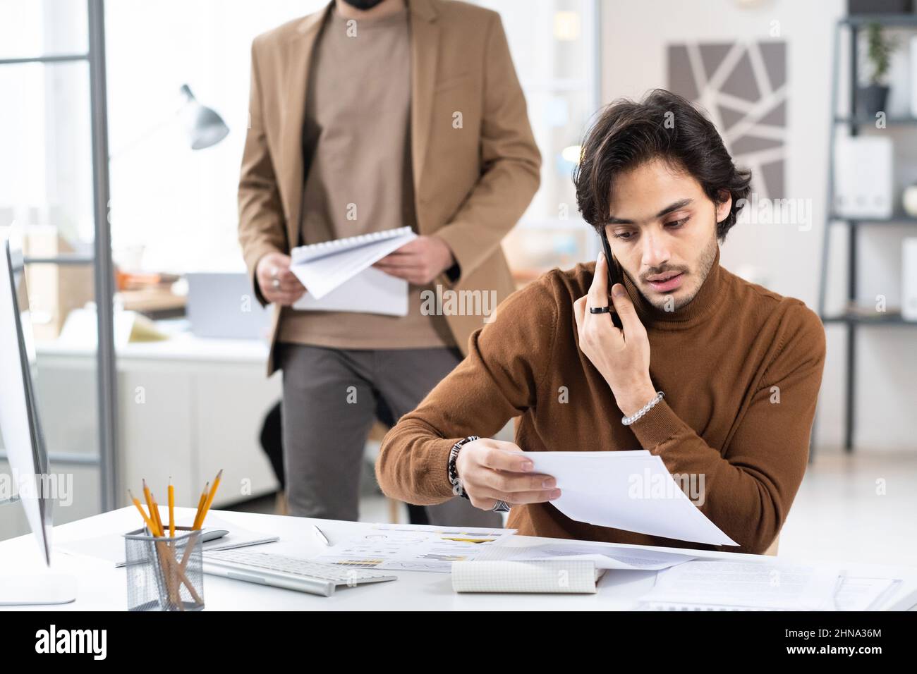 Giovane uomo d'affari che legge contratto mentre ha una conversazione sul telefono mobile, lui lavora al suo posto di lavoro in ufficio Foto Stock