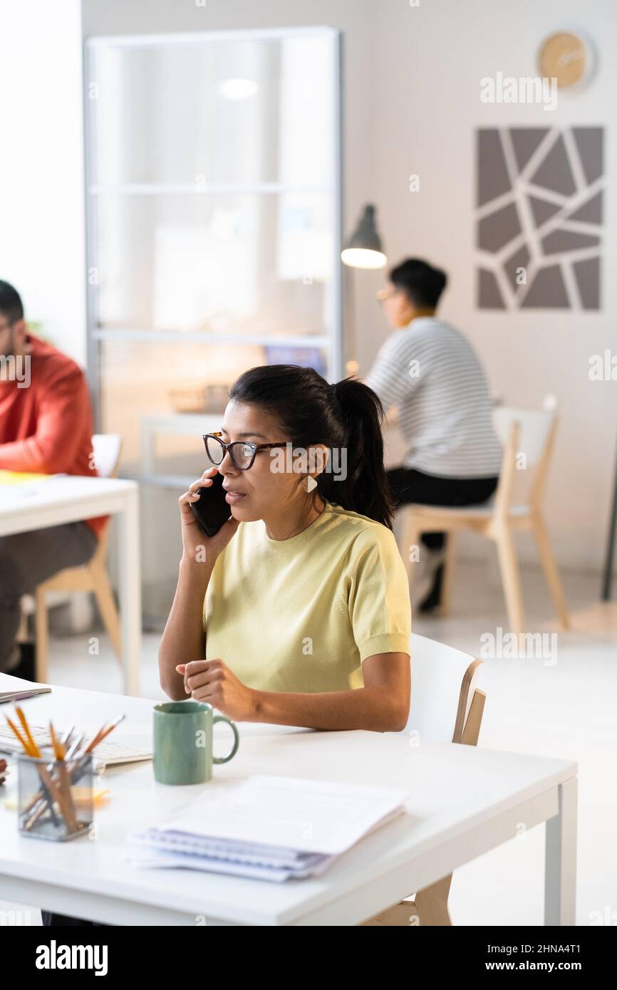Donna d'affari in occhiali concentrandosi sulla chiamata seria sul telefono mobile mentre si siede al suo posto di lavoro in ufficio Foto Stock