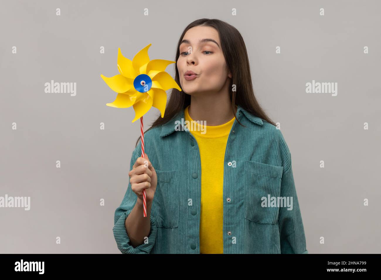 Mulino a mano Ritratto di felice bella giovane donna adulta soffiando a carta mulino a vento, ruota a pinneheel giocattolo sul bastone, indossando casual stile giacca. Studio interno girato isolato su sfondo grigio. Foto Stock