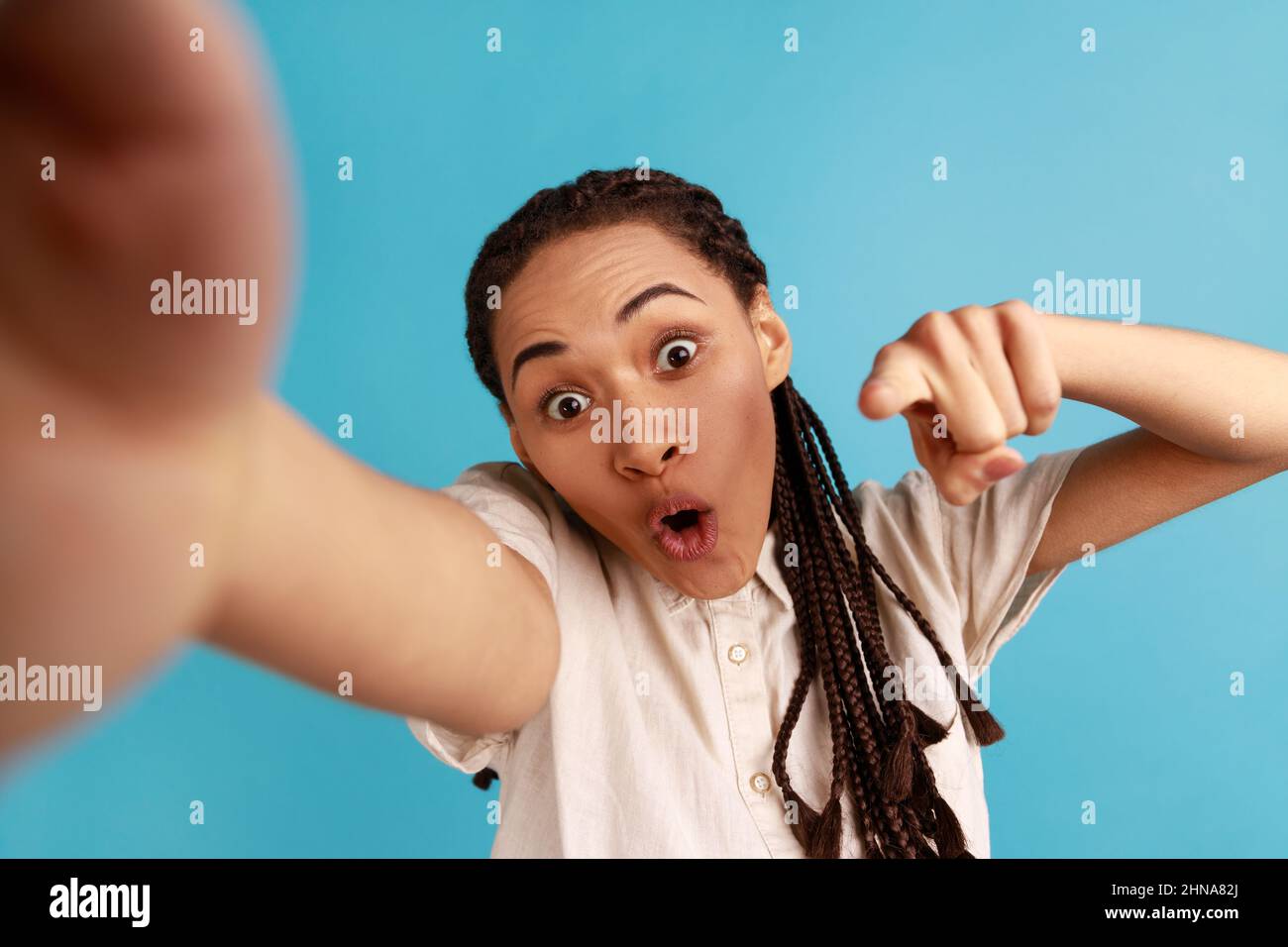 Scioccata donna con dreadlocks nero ha stupito l'espressione del volto, facendo selfie, esprime emozioni stupite, puntando il dito alla telecamera, POV. Studio interno girato isolato su sfondo blu. Foto Stock