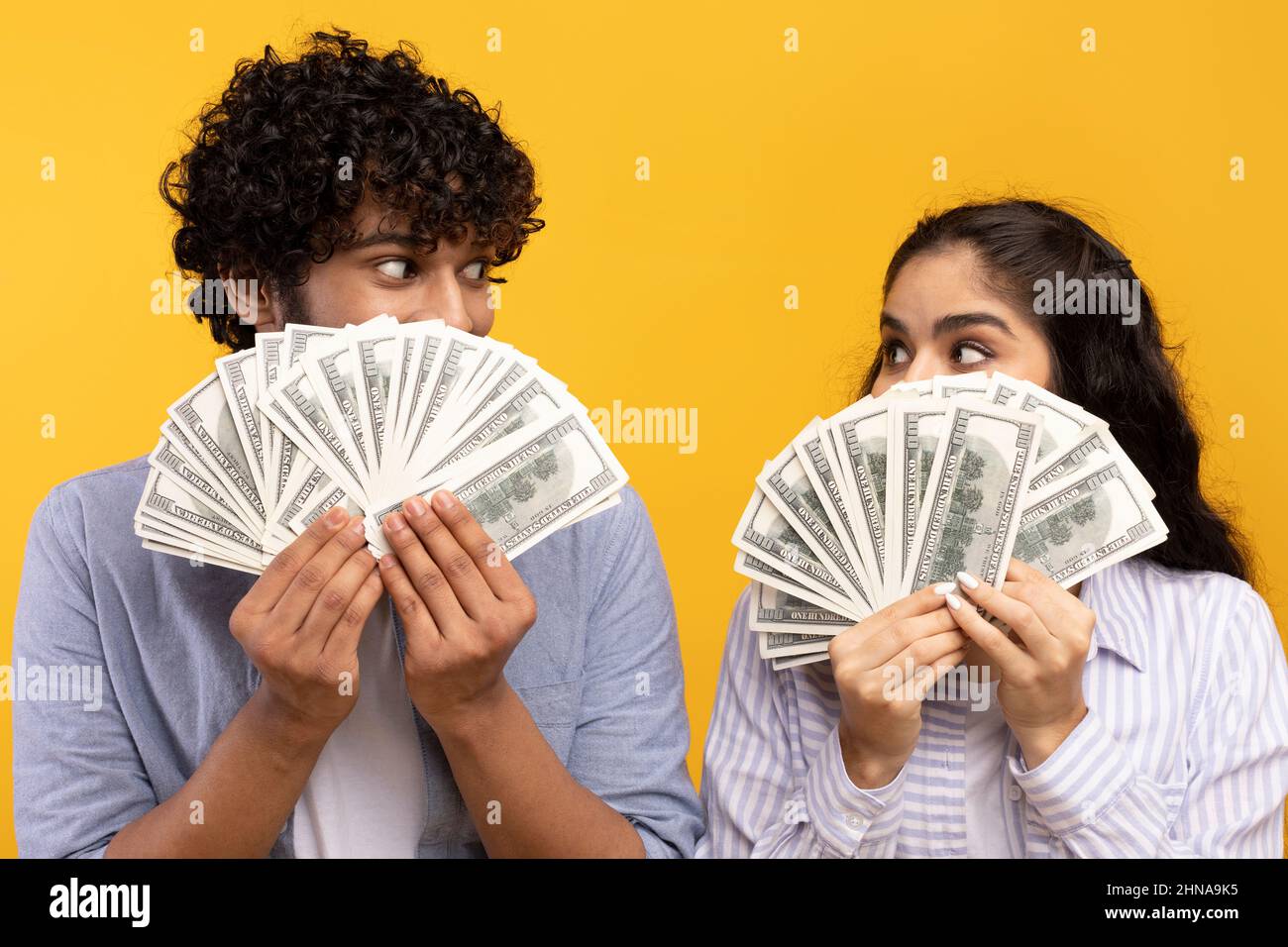 Concetto di grande successo. Giovane coppia indiana che tiene e copre le facce con un sacco di dollari ventilatore di cassa, sfondo giallo studio Foto Stock