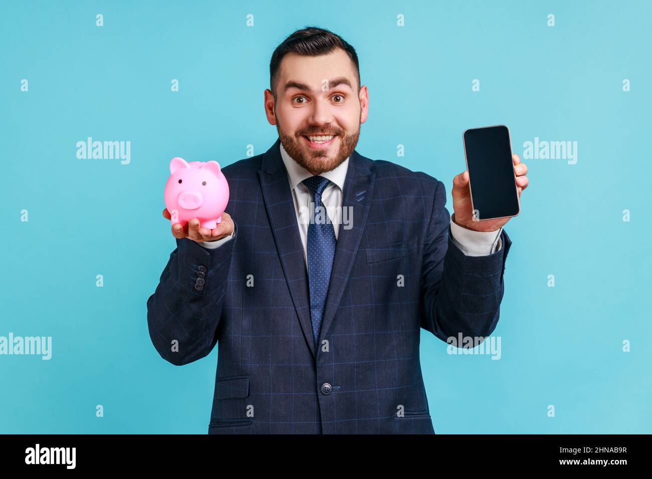 Uomo sorridente felice con barba indossando un abito ufficiale con smartphone con schermo vuoto e pig bank, cashback, portafoglio online. Studio interno girato isolato su sfondo blu. Foto Stock