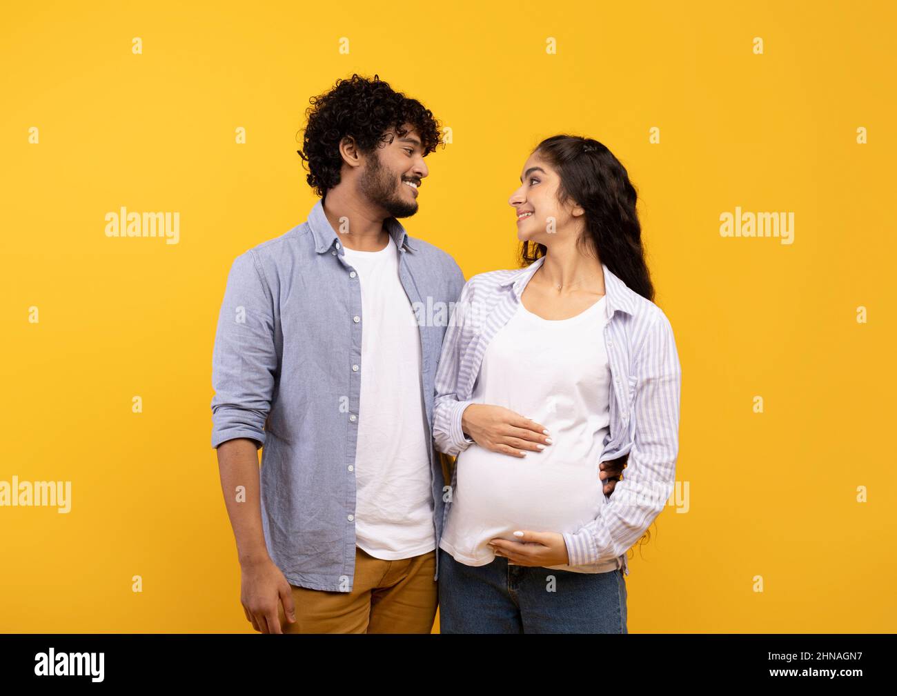 Coppia indiana felice che aspetta il bambino, posando su sfondo giallo, abbracciandosi e guardandosi l'un l'altro Foto Stock