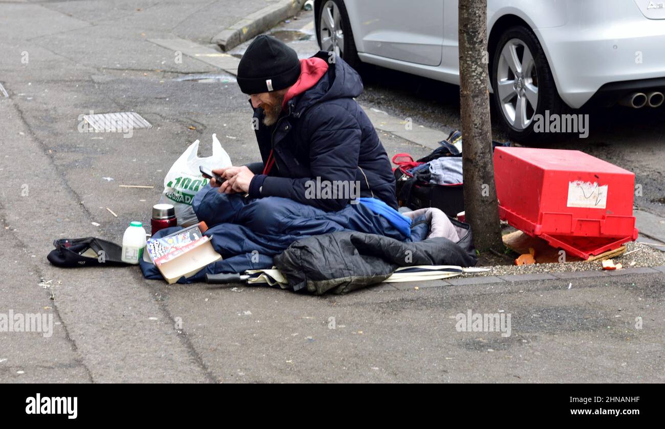 Uomo seduto sul marciapiede con accoccolato in cappotto pesante con sacchi a pelo e altre posizioni Foto Stock