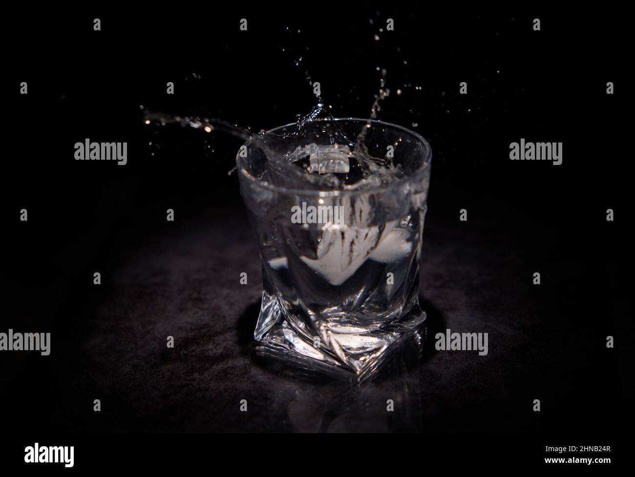 Spruzzi d'acqua da un pezzo di ghiaccio che cadono in un vetro trasparente con sfondo nero Foto Stock