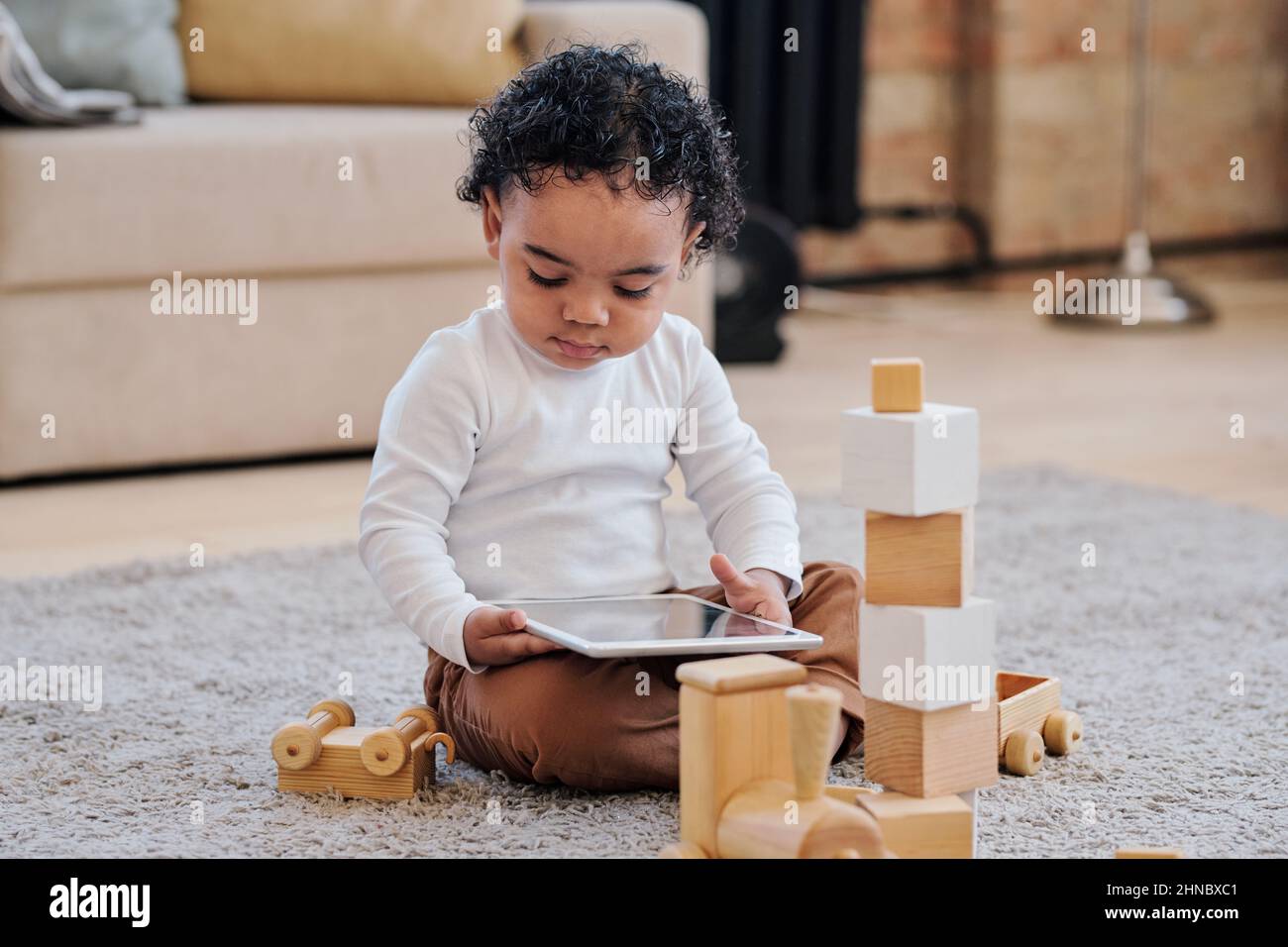 Serio curioso ragazzo afro-americano bambino in camicia bianca seduta sul pavimento tra giocattoli di legno e utilizzando tablet Foto Stock