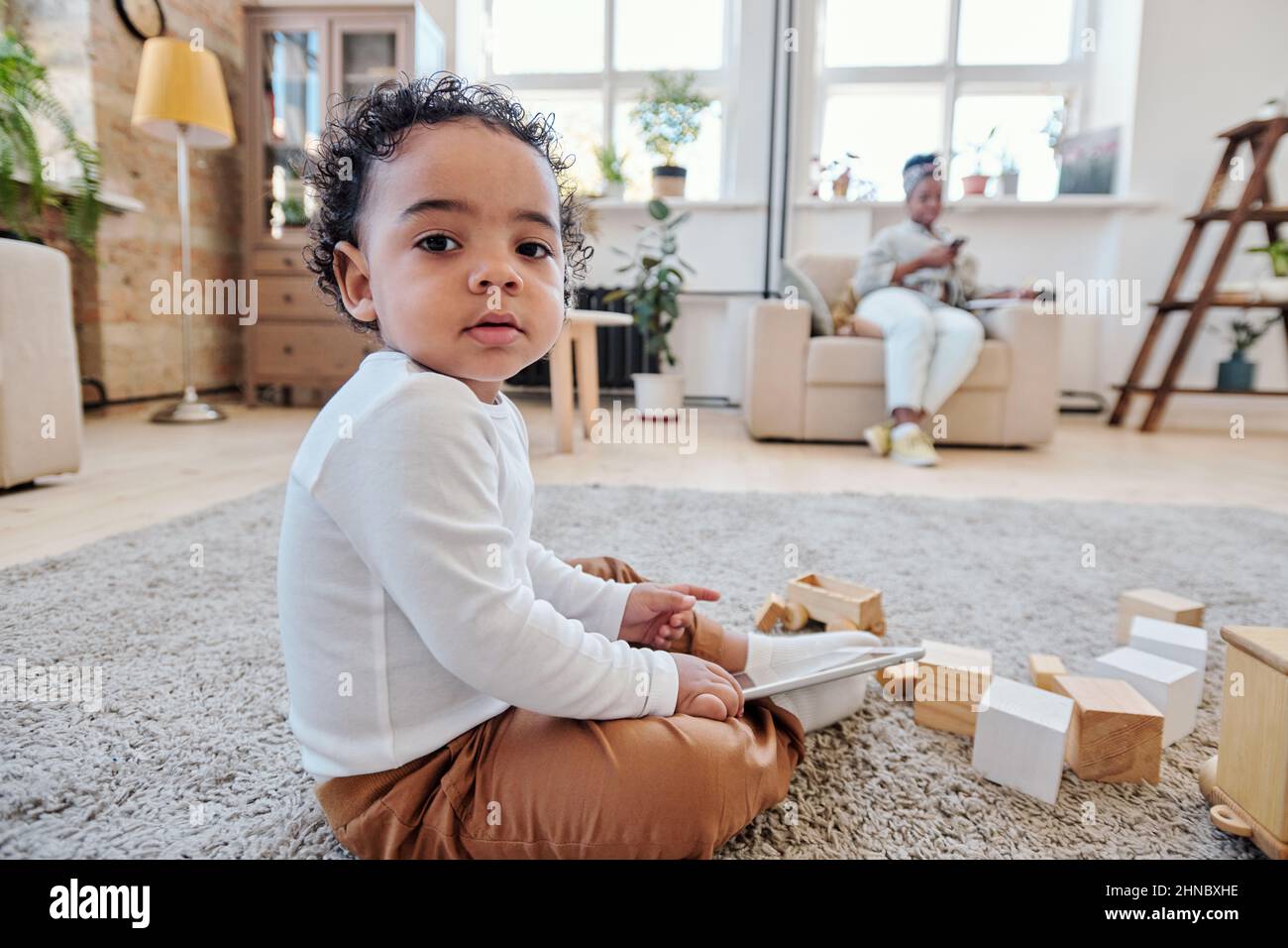 Ritratto di ragazzo afroamericano serio carino seduto sul pavimento con giocattoli di legno e tenere tablet Foto Stock