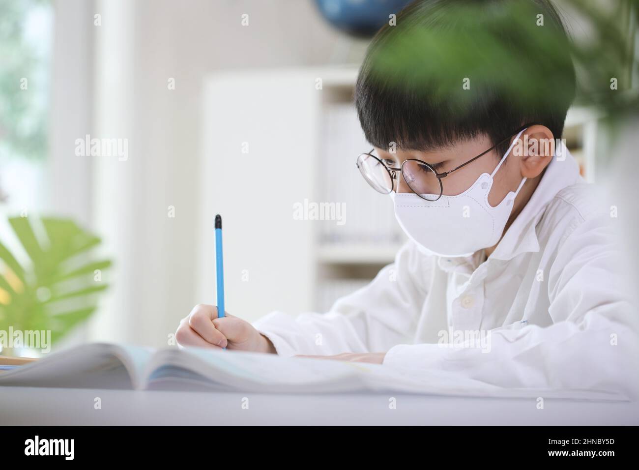 Studente intelligente e carino in una maschera che studia duro mentre si concentra sta imparando mentre prende appunti in libri e note alla sua scrivania. Foto Stock