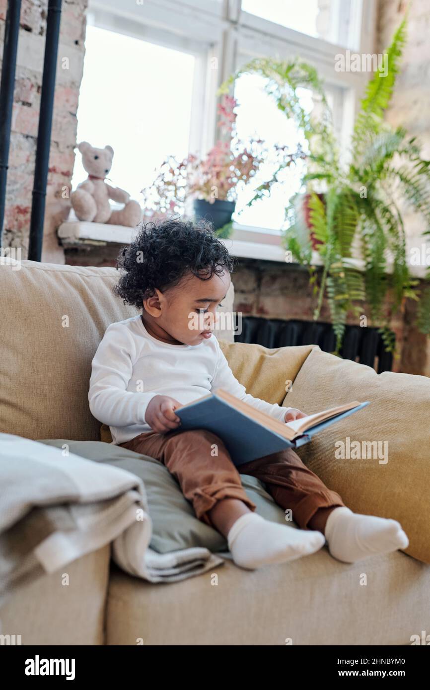 Concentrato curioso afroamericano toddler seduta con libro in poltrona e lettura in soggiorno Foto Stock