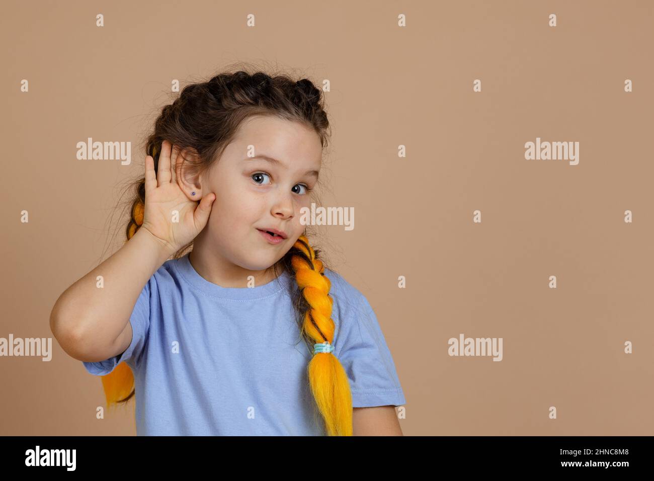 Curioso interessato piccola ragazza caucasica che hanno trecce kanekalon con occhi luminosi che tengono le orecchie per ascoltare qualcosa che guarda la macchina fotografica sul beige Foto Stock