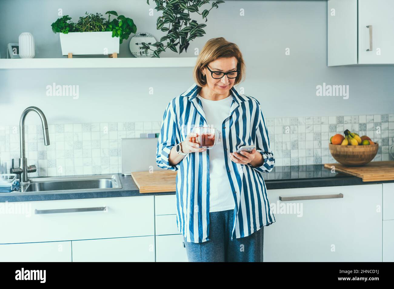 Donna sorridente di mezza età che guarda il suo telefono cellulare e beve il tè mentre si trova nella cucina moderna. Abitudini e rituali del mattino. Online addicti Foto Stock