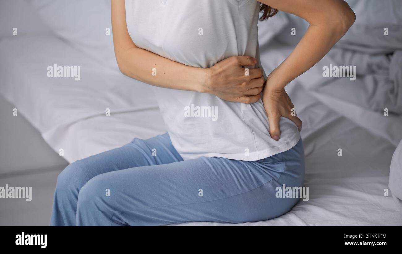 vista parziale della donna in pigiama che tocca il lombo doloroso mentre si siede a letto Foto Stock