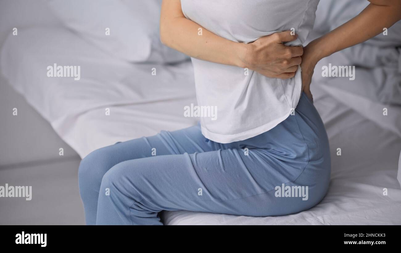 vista parziale della donna seduta a letto in pigiama e sofferente di dolore addominale Foto Stock