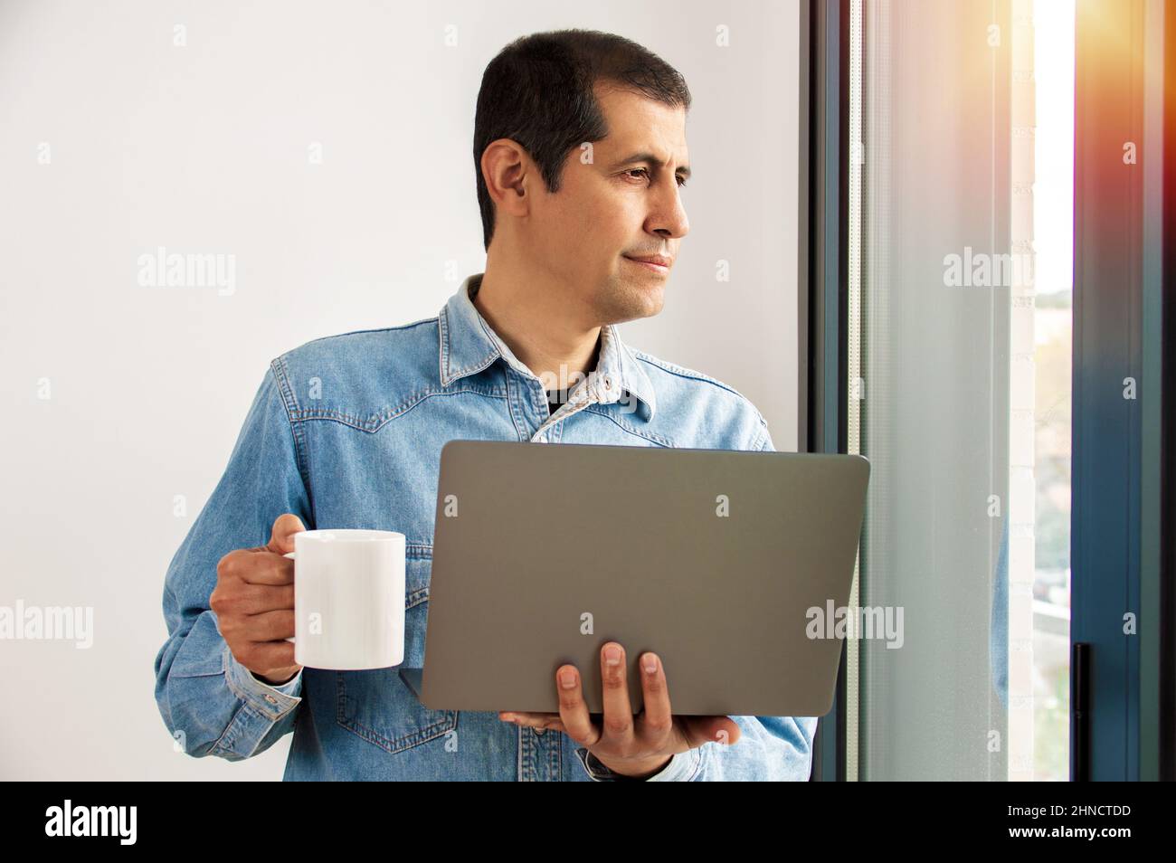 Scatto di un bel giovane uomo d'affari che beve caffè mentre lavora su un laptop nel suo ufficio domestico e guarda fuori dalla finestra Foto Stock