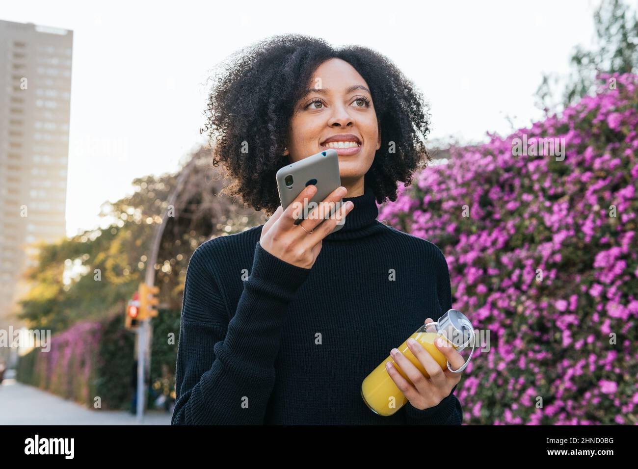Positiva femmina afroamericana con succo d'arancia guardando in su e registrare il messaggio audio sullo smartphone mentre si trova vicino a cespugli fioriti con porpora Foto Stock