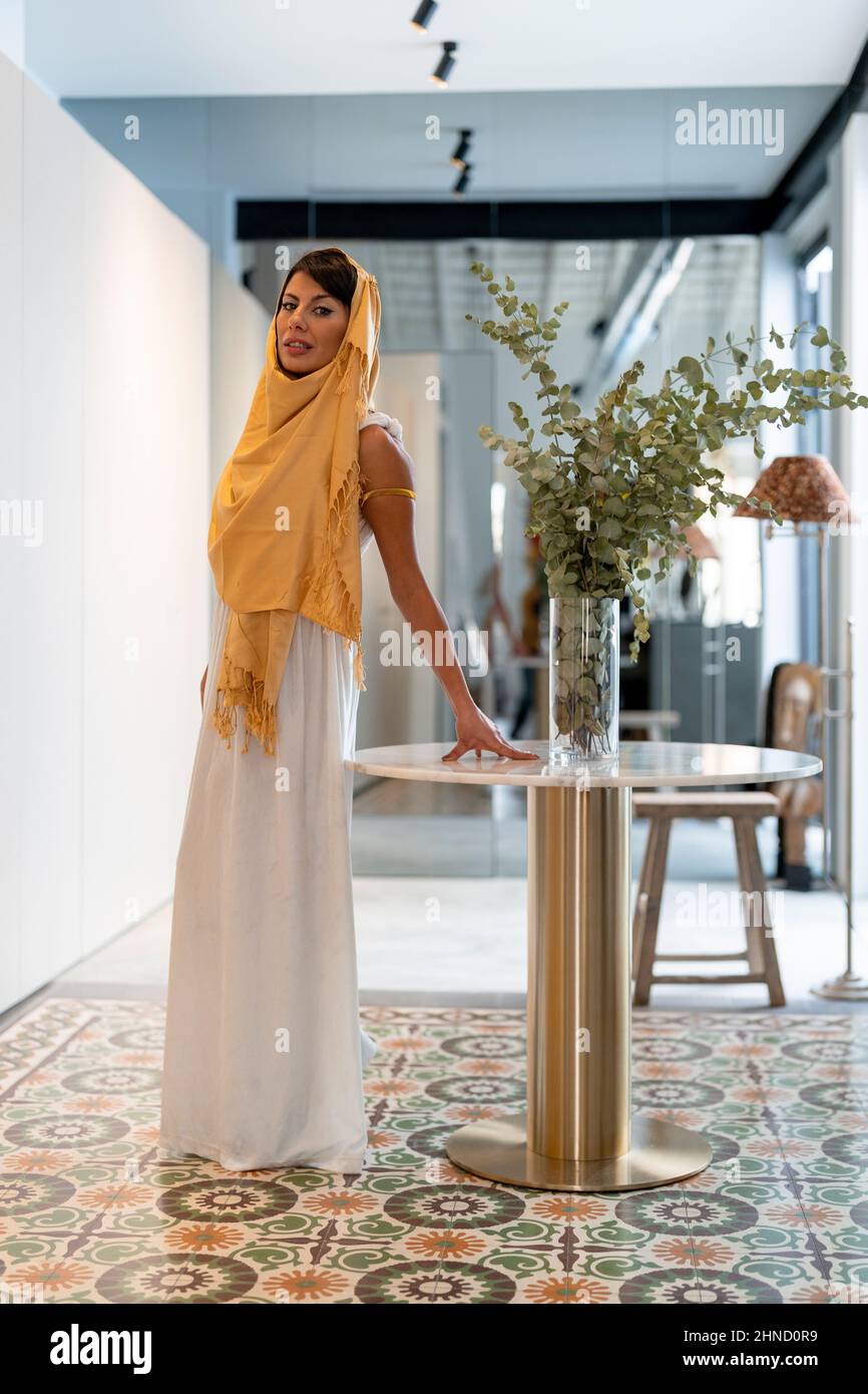 Corpo pieno di affascinante femmina musulmana in hijab tradizionale guardando la macchina fotografica mentre si trova vicino al tavolo con fiori in camera Foto Stock