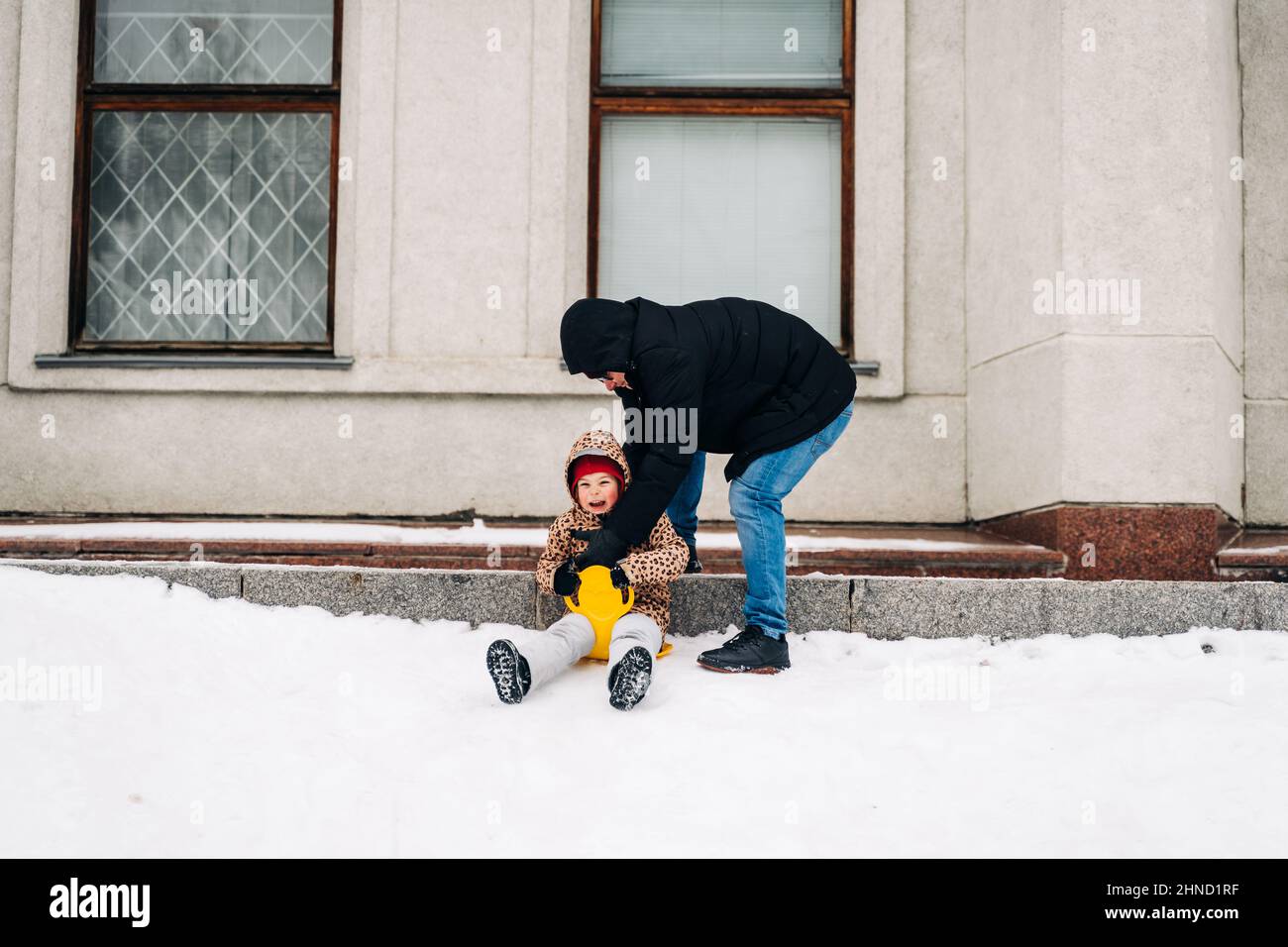Corpo pieno di padre premuroso che aiuta il bambino su outerwear a sedersi sul piatto giallo della neve nella fredda giornata invernale in città Foto Stock