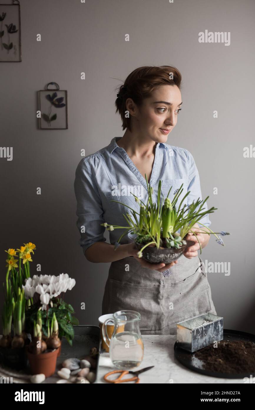 Contenuto donna giardiniere in grembiule in piedi con assortiti vasi di fiori freschi e piante a tavola a casa e guardando via Foto Stock
