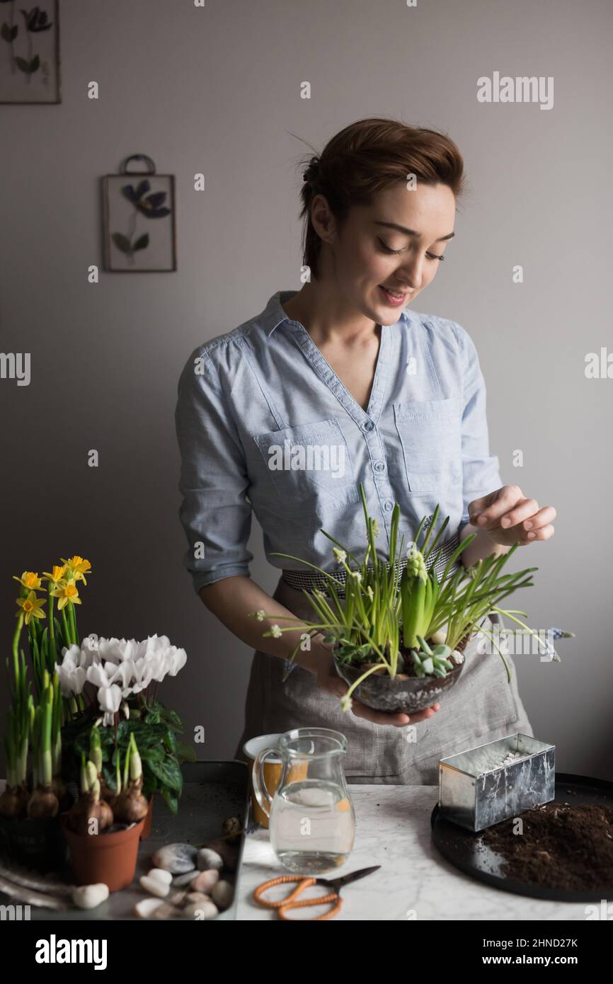 Contenuto donna giardiniere in grembiule in piedi con assortiti vasi di fiori freschi e piante a tavola a casa e guardando verso il basso Foto Stock