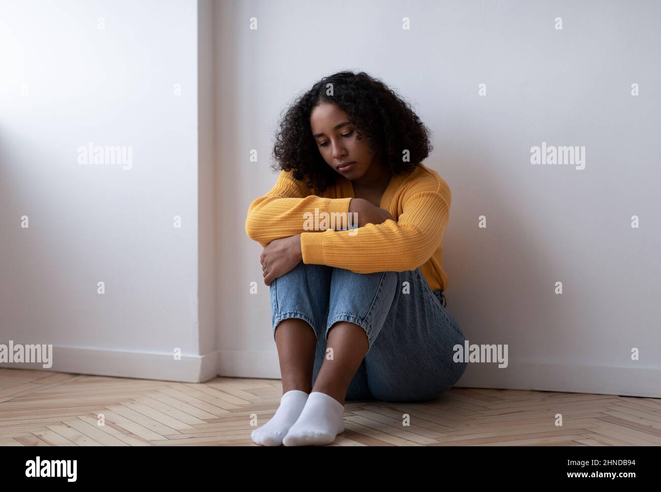 Tutta la lunghezza di giovane donna nera che soffre di depressione, seduta sul pavimento e abbracciando le ginocchia a casa, spazio copia Foto Stock
