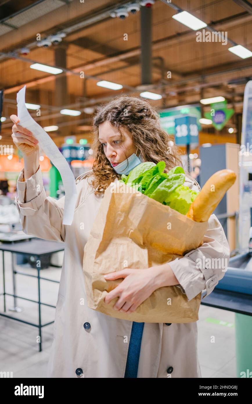 La giovane donna è sorpresa spiacevolmente mentre osserva in un controllo della carta in un supermercato della drogheria e tiene una borsa della spesa. Aumento dei prezzi dei prodotti alimentari. Foto Stock