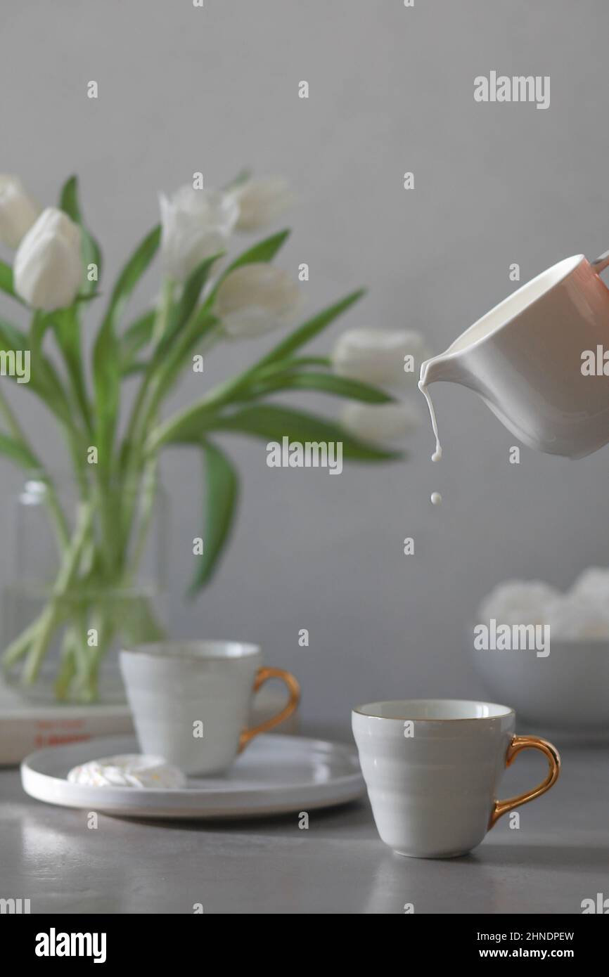 Versare il latte in una tazza con caffè, un mazzetto di tulipani bianchi su sfondo grigio. Colazione Foto Stock