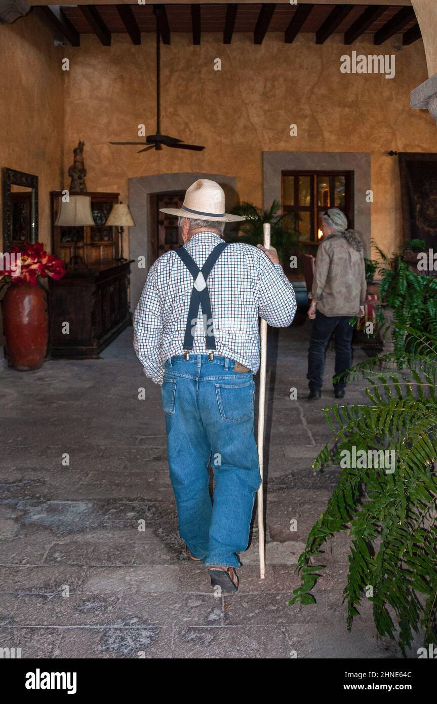 Semplice e folcloristico uomo anziano con jeans blu, bretelle e cappello di paglia passeggia con un bastone attraverso un'elegante hall in stile spagnolo in Messico. Foto Stock