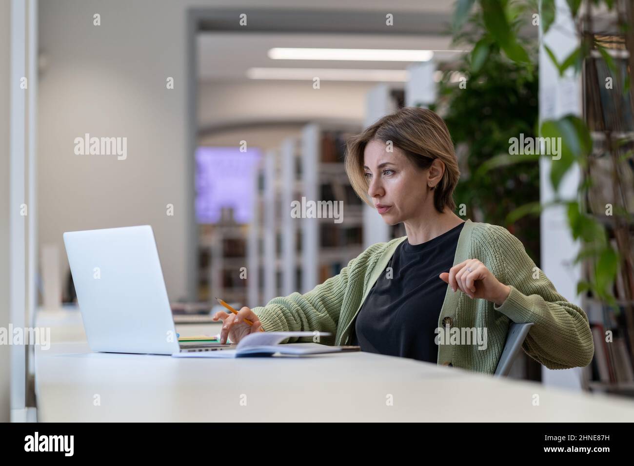 Insegnante di università femminile focalizzata e pensiva che si prepara a una conferenza online su laptop nella moderna biblioteca Foto Stock