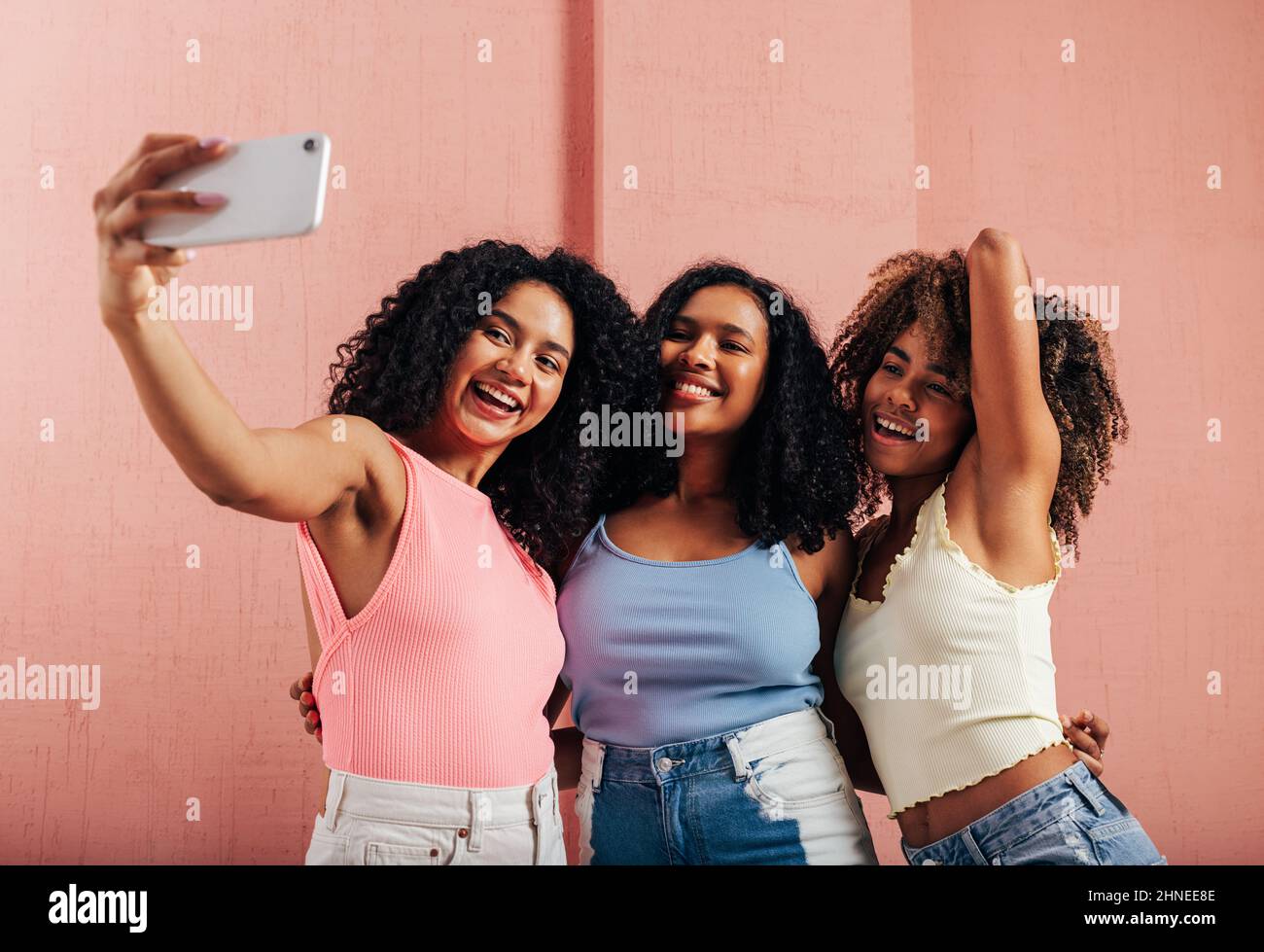 Tre donne felici con capelli ricci che prendono selfie contro la parete rosa Foto Stock