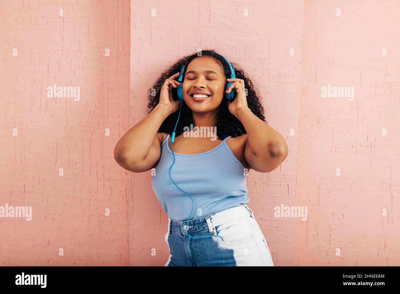 Donna che indossa cuffie blu per ascoltare la musica mentre si alza con gli occhi vicini alla parete rosa Foto Stock