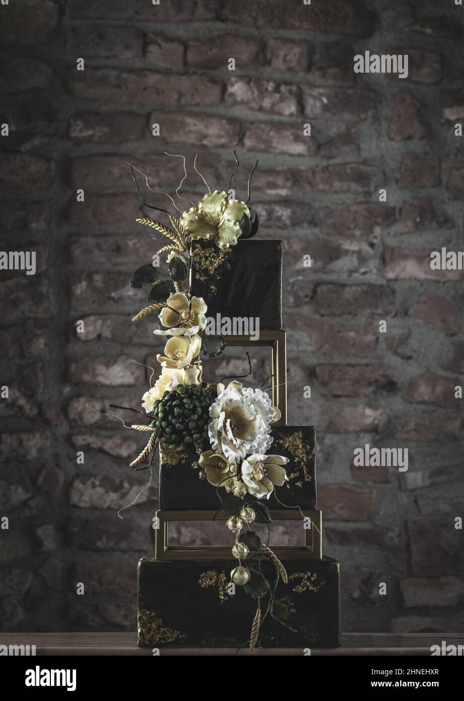 Torta nuziale quadrata a tre piani con fiori e accenti di verde scuro su sfondo di pietra Foto Stock