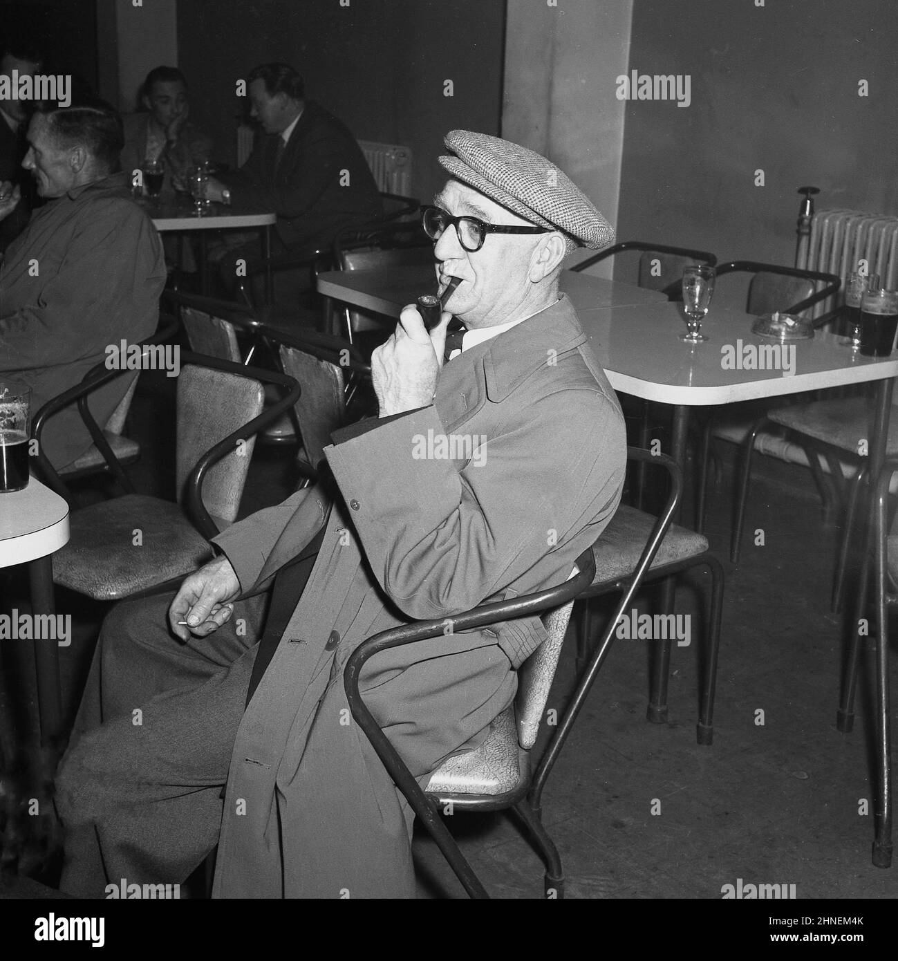 1950s, storico, un uomo di lavoro che indossa giacca e pantaloni, un impermeabile, cappotto e fumare un tubo, seduto a bere una bevanda, una pinta di birra, nel sociale società dopo il lavoro, Port Talbot, Galles, Regno Unito. Foto Stock
