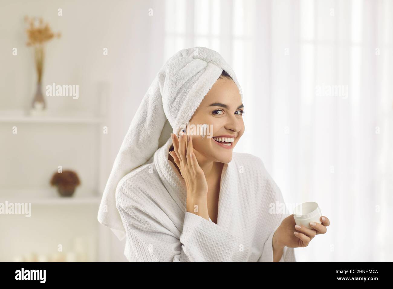 Sorridendo bella giovane donna che applica crema facciale mentre fa la sua routine di cura della pelle Foto Stock