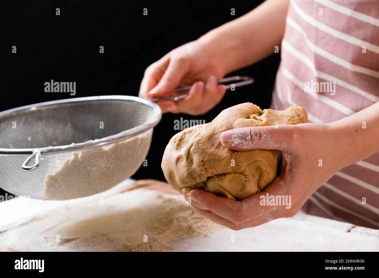 Primo piano la donna setaccia la farina di grano attraverso un setaccio in un recipiente di miscelazione per la preparazione dell'impasto per la torta e la panetteria su sfondo nero. Impastare l'impasto per ottenere la pasta fatta in casa. Foto Stock