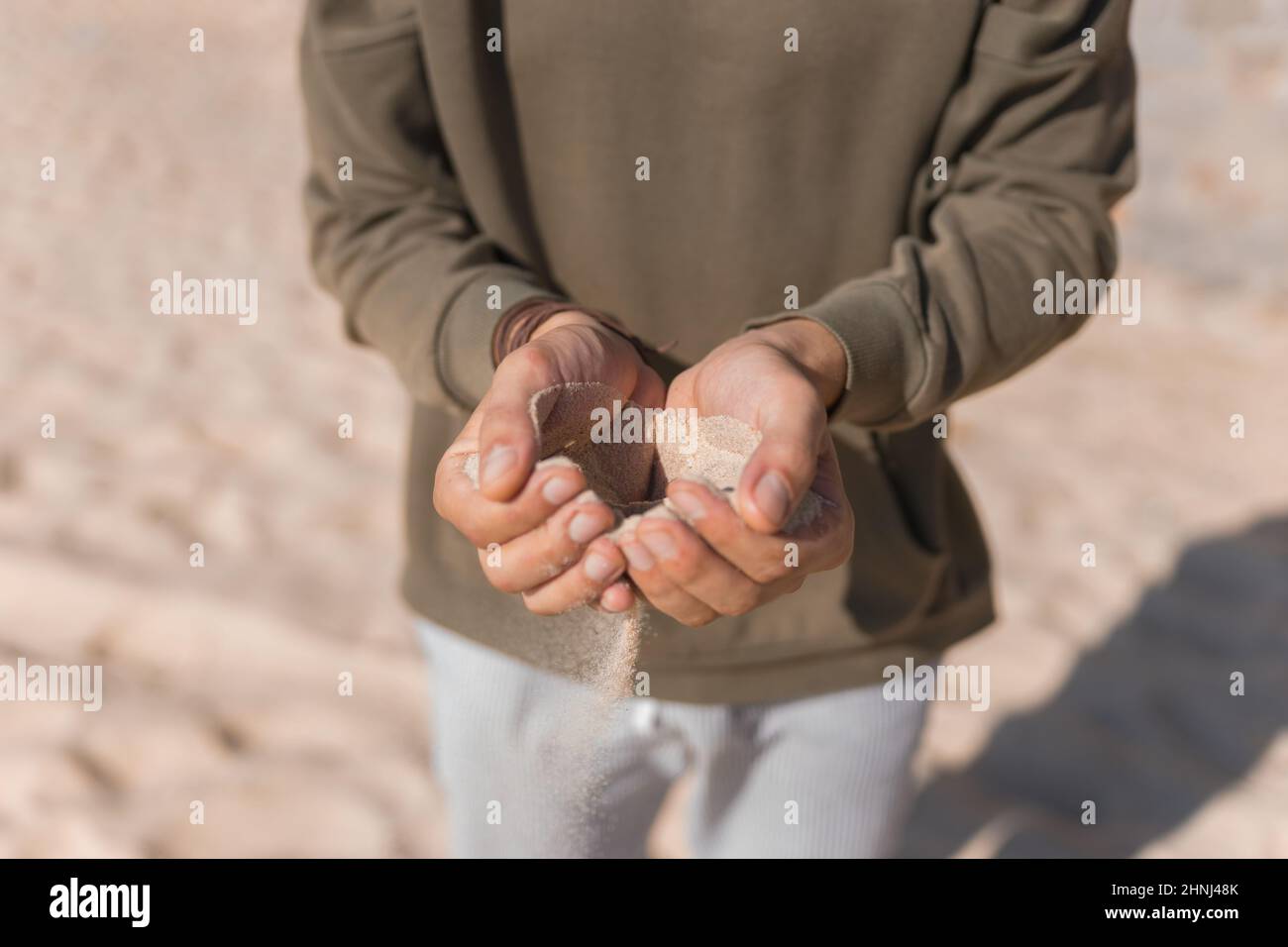 Uomo che tiene la sabbia bianca nelle sue mani. Palme maschili con la sabbia sulla spiaggia. Versare sabbia. Felpa con cappuccio casual. Foto di alta qualità Foto Stock