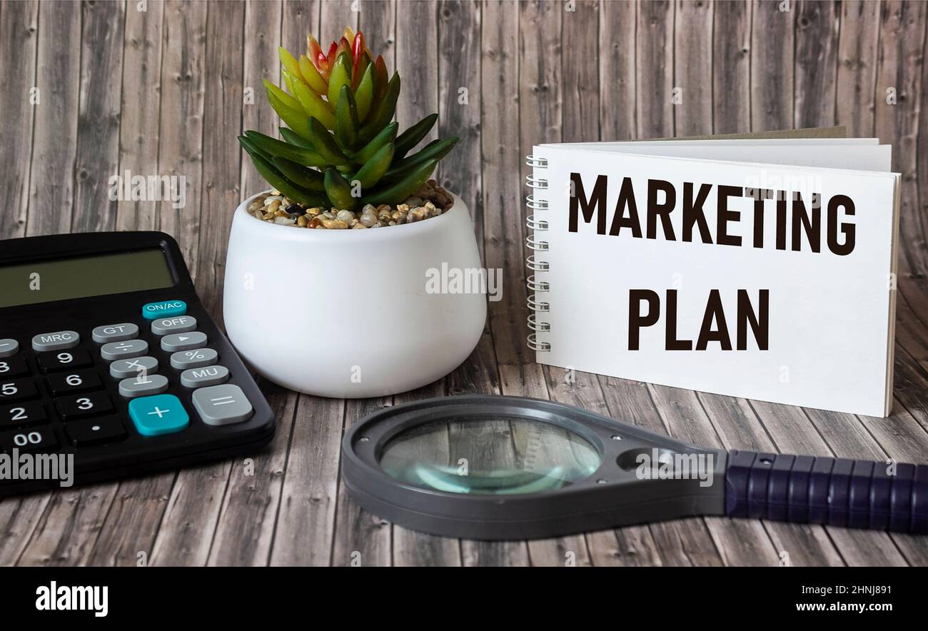 Piano di marketing scritto in un blocco note, un tavolo di legno con una calcolatrice, una lente d'ingrandimento e una penna. Immagine di concetto per l'intestazione del blog o l'immagine dell'intestazione. Foto Stock