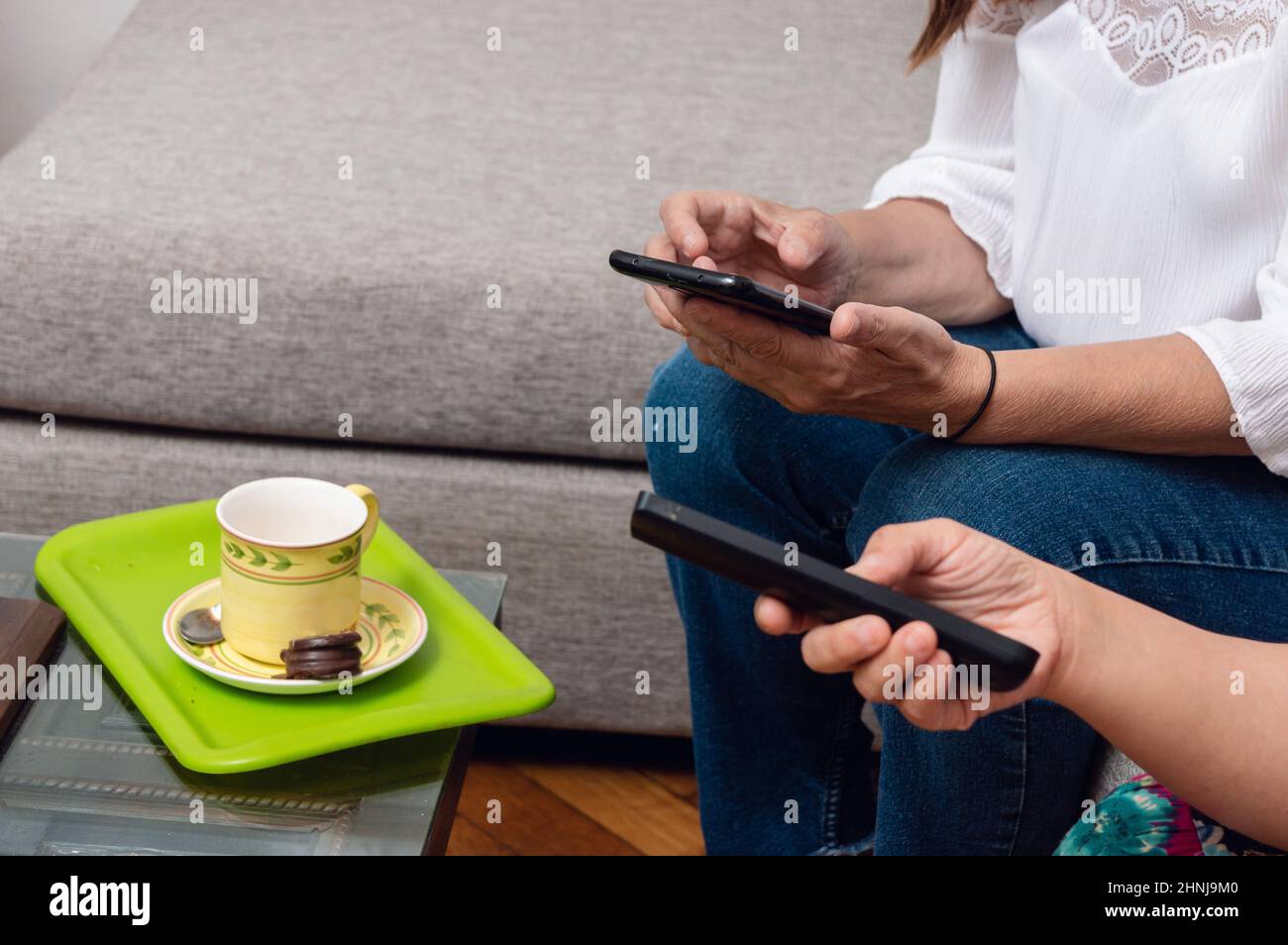 primo piano di mani caucasiche femminili in un soggiorno appartamento con un telefono e un televisore, con una tazza di caffè e un cioccolato alfa Foto Stock