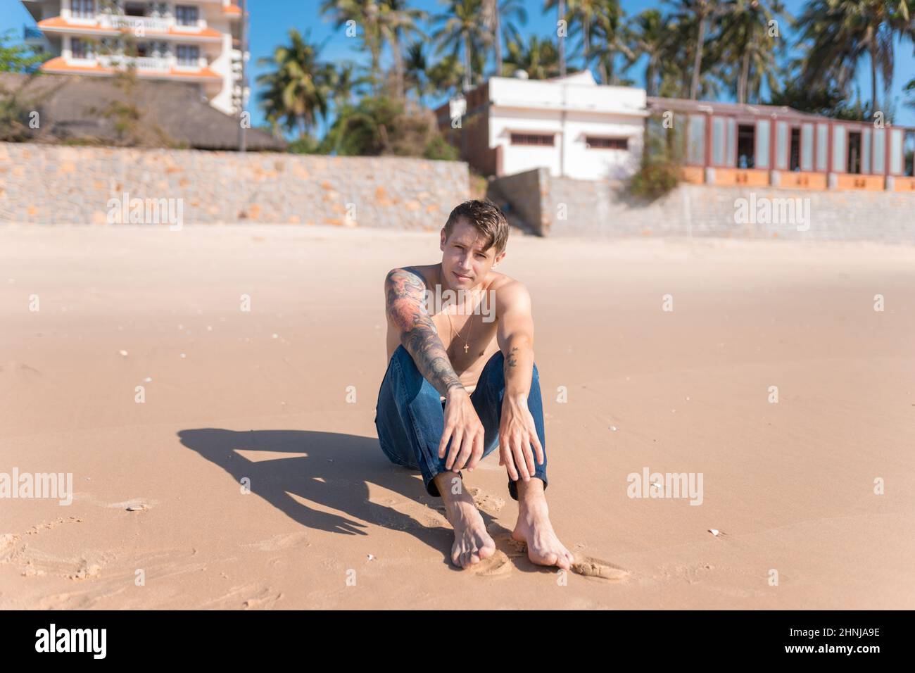 Attraente giovane caucasico nudo-chested uomo con tatuaggio full-ARM seduto sulla sabbia sulla spiaggia. Relax sulla spiaggia. Foto di alta qualità Foto Stock