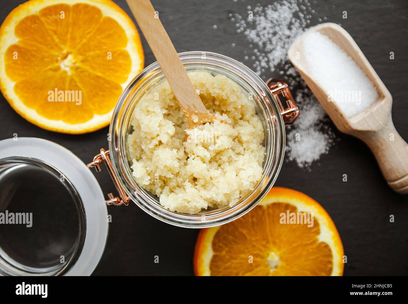 Scrub corpo di zucchero fatto in casa in vaso di vetro, decorato con fette di arancia fresca e cucchiaio di legno con polvere di zucchero sul tagliere di pietra nera. Foto Stock