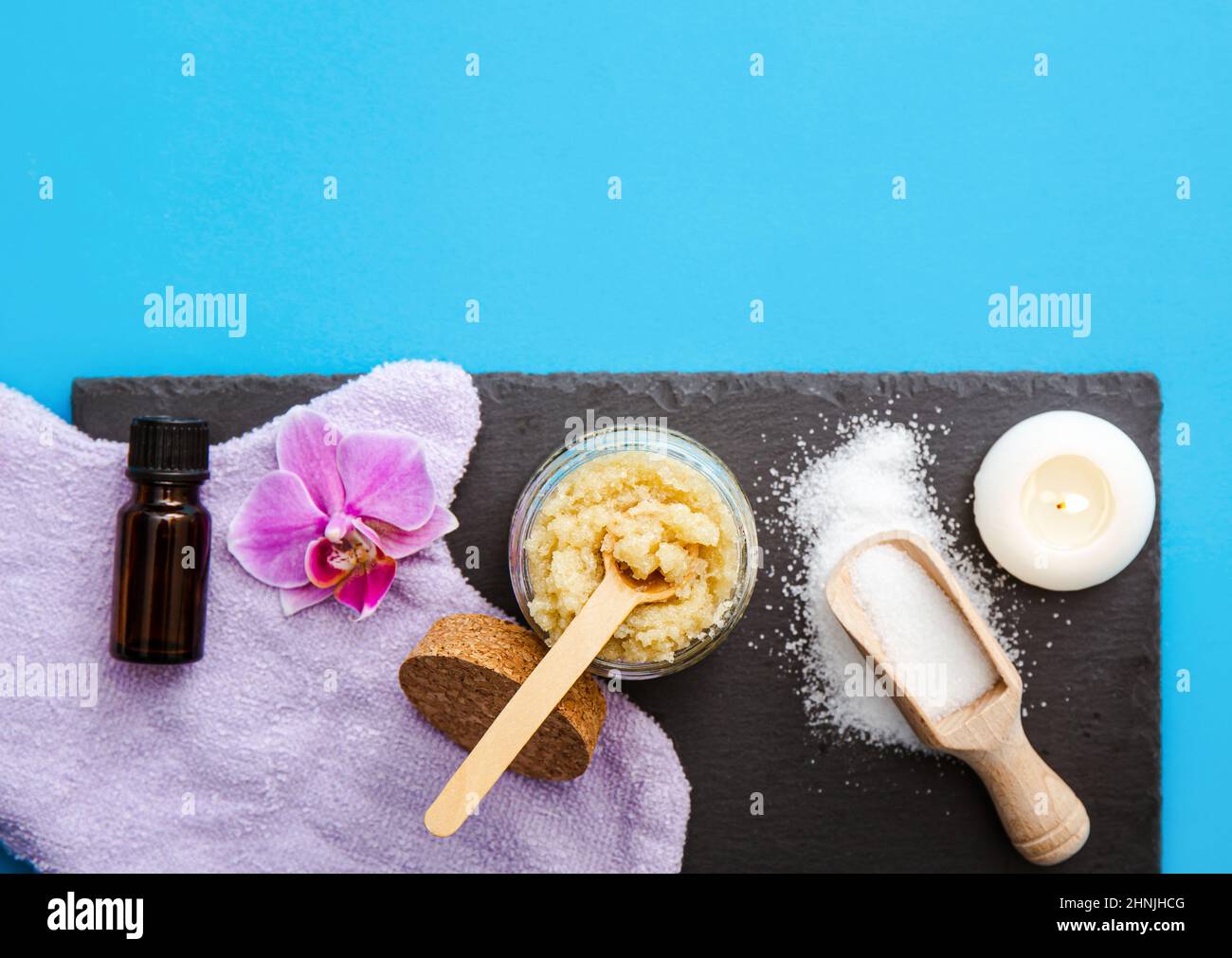 Scrub corpo di zucchero fatto in casa in vaso di vetro con ingredienti di scrub fatti in casa, zucchero e olio aromatico, olio essenziale. Candela che brucia e fiore rosa orchidea. Foto Stock