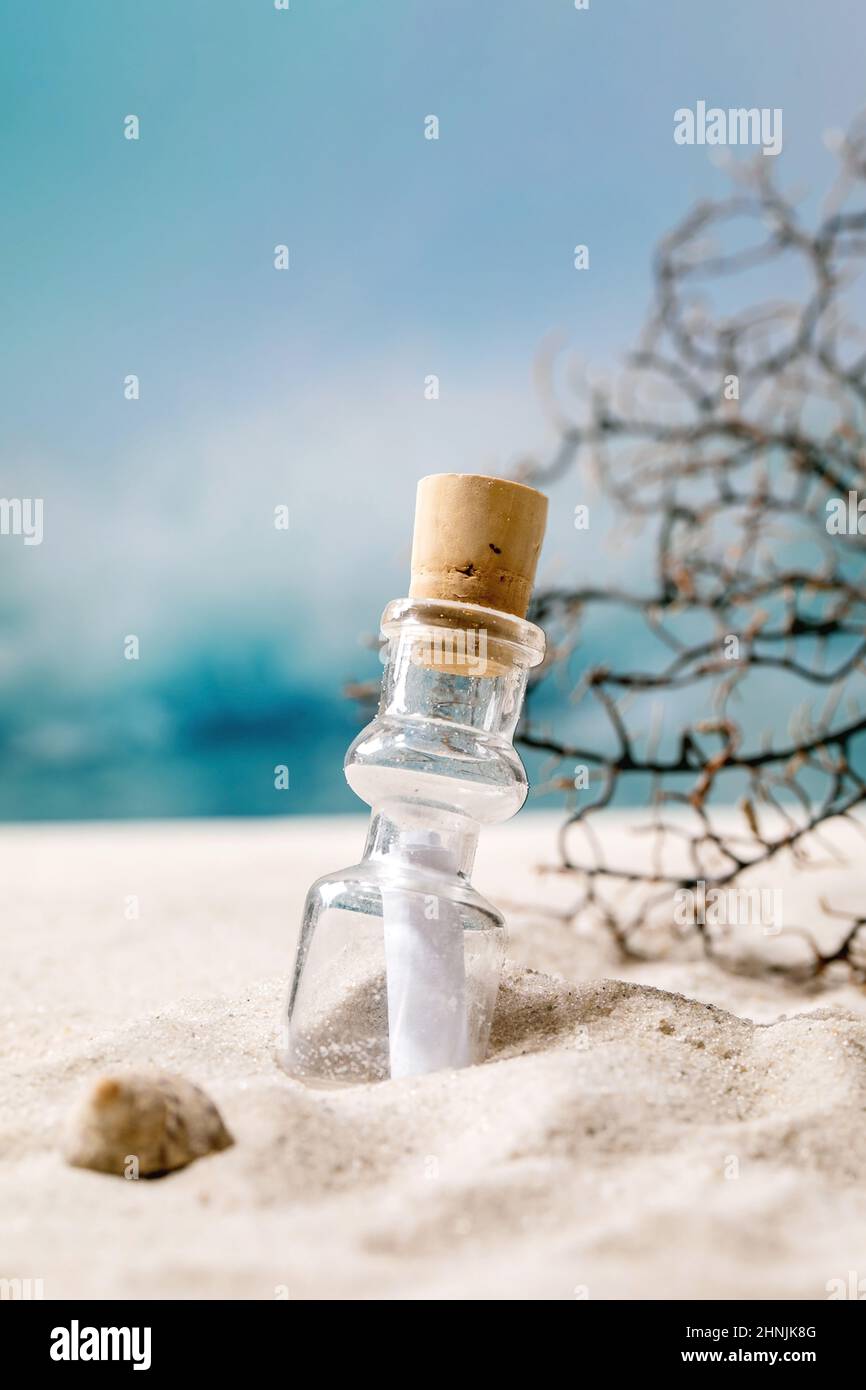 Bottiglia di vetro con carta per messaggi all'interno sulla spiaggia di sabbia marina estiva con onde e ramo di corallo sullo sfondo. Aiuto SOS concetto di messaggio destino. Foto Stock