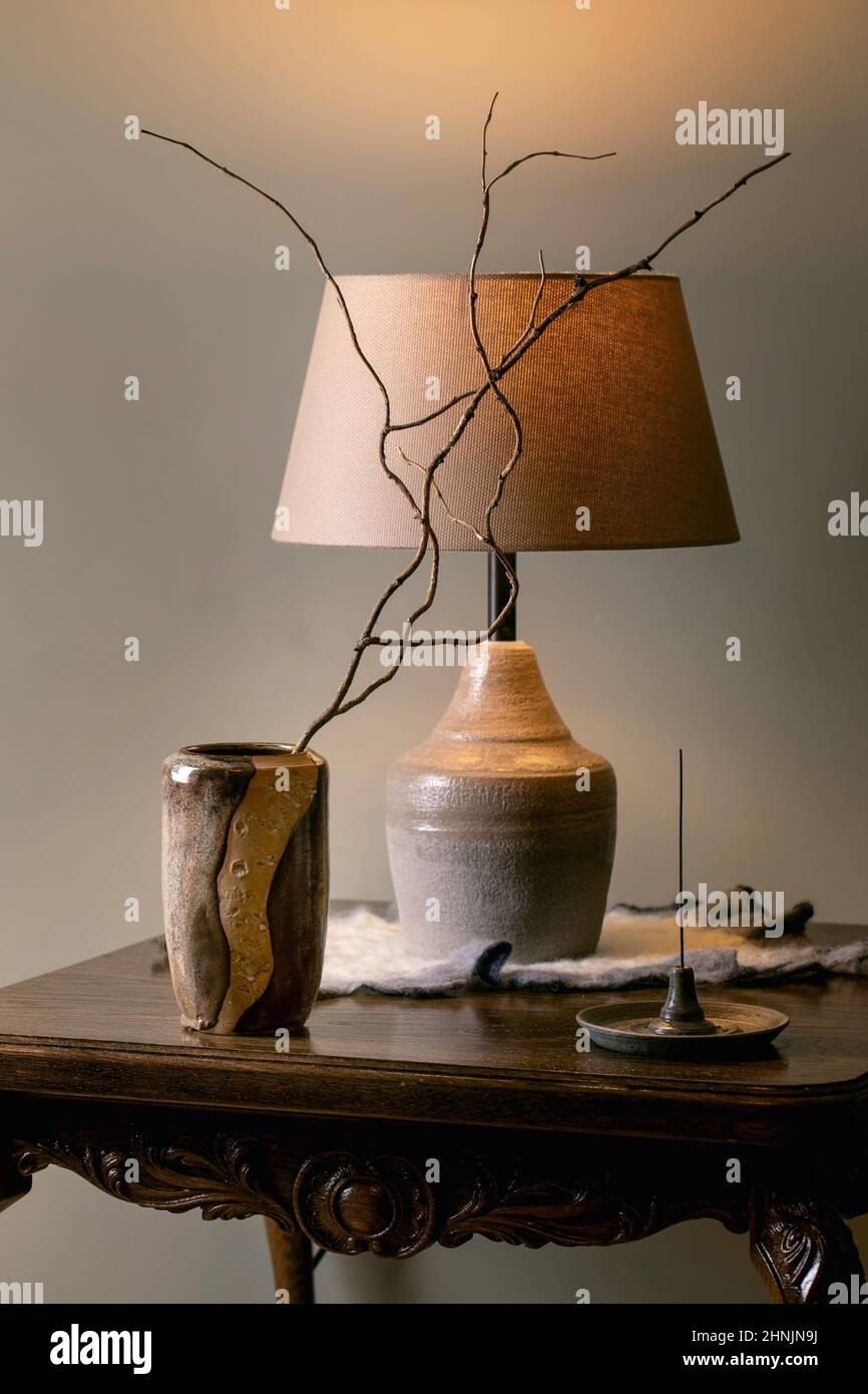 Interni domestici. Ramoscelli a secco in vaso in ceramica artigianale beige, supporto per incenso, lampada da tavolo in ceramica posta su un antico tavolo in legno con parete grigia Foto Stock