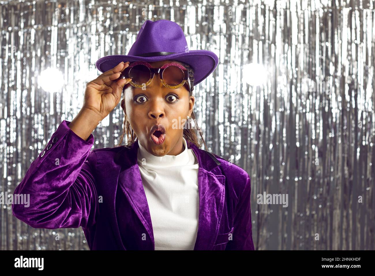 Donna con pelle scura e giacca in corduroy ti guarda a sorpresa su un lucido sfondo di festa in lamina. Foto Stock