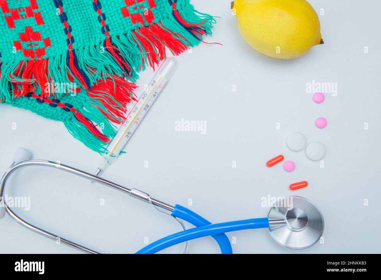 Una calda sciarpa invernale, un termometro, un limone, uno stetoscopio e pillole con capsule giacciono su sfondo bianco. Foto Stock