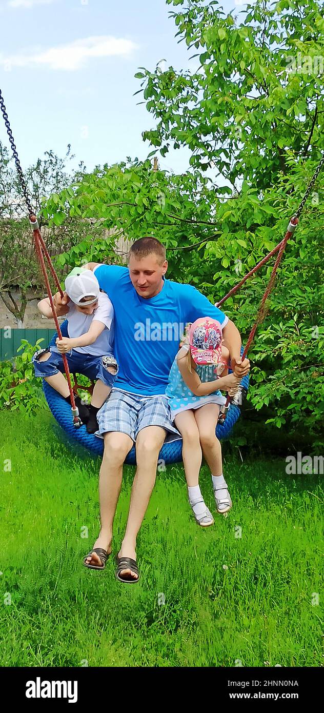 Buon padre con bambini che cavalcano l'altalena in giardino. La famiglia gode l'estate Foto Stock