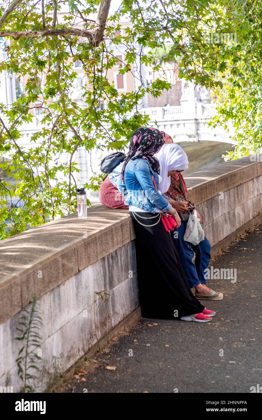 Le donne muscolose con sciarpa si godono la passeggiata lungo il Tevere e utilizzare il cellulare per inviare messaggi a Roma Foto Stock